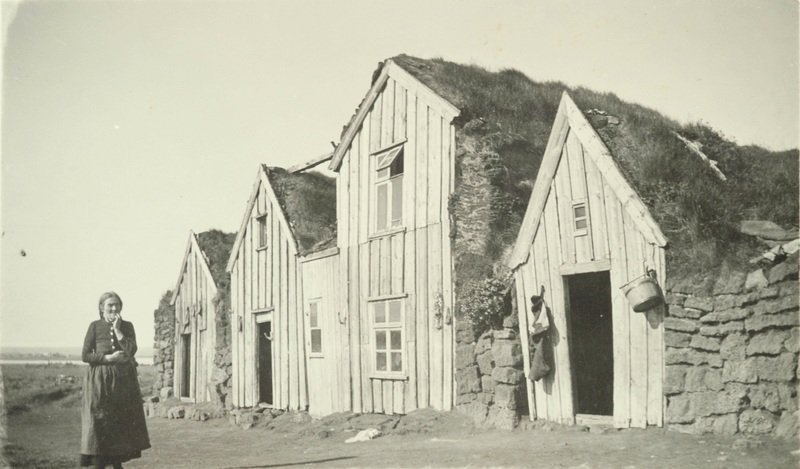 Torfbær með fjórum burstum. Fyrir framan bæinn stendur kona sem styður hendi undir kinn. Myndin er tekin á árunum 1925-1935. Ljósm. Þjóðminjasafn Íslands.