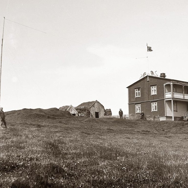 „Málmeyjarhúsið 25. júní 1931. Hér er húsið 6 ára gamalt og í sínu besta standi. Hluti gamla bæjarins stendur enn ofan við og fjósbyggingin komin norðan undir. Búið er að draga fána að húni, líklega í tilefni myndatökunnar eða komu Arthurs Gook sem hefur gætt þess að útvarpsstöngin nyti sín á myndinni, enda upphaflega reist 1928 vegna útvarpsstöðvar hans. Þessi stöng virðist ætla að standa í heila öld og er enn árið 2020, það eina sem upp úr gnæfir á bæjarstæðinu. Einn fóstursonurinn er við stöngina en annar við húsgaflinn og líklega er það Gísli Konráðsson meðeigandi í Málmey sem ber við loft á myndinni. Hrúgaldið sunnan við húsið og grindverkið mun vera bænhústóftin og mótar vel fyrir bænhúsgarðinum í hálfhring utan um hana,“ segir Hjalti Pálsson, Byggðasöguritari, í myndatexta.  MYND ÚR X. BINDI BYGGÐASGÖU SKAGFIRÐINGA